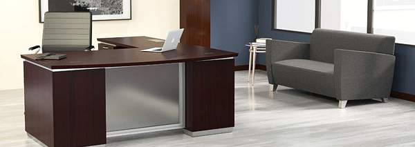 Dark mahogany L-shaped office desk angled by a window, with a grey lounge sofa opposite in a two-zone office layout
